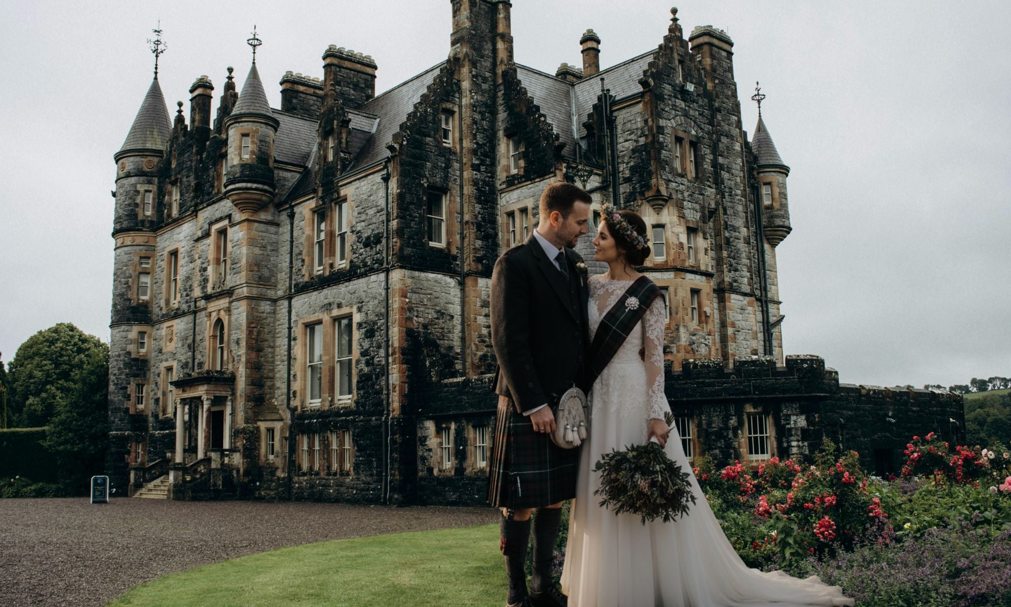 Couple in Scottish attire standing in front of a large castle with a cloudy sky.