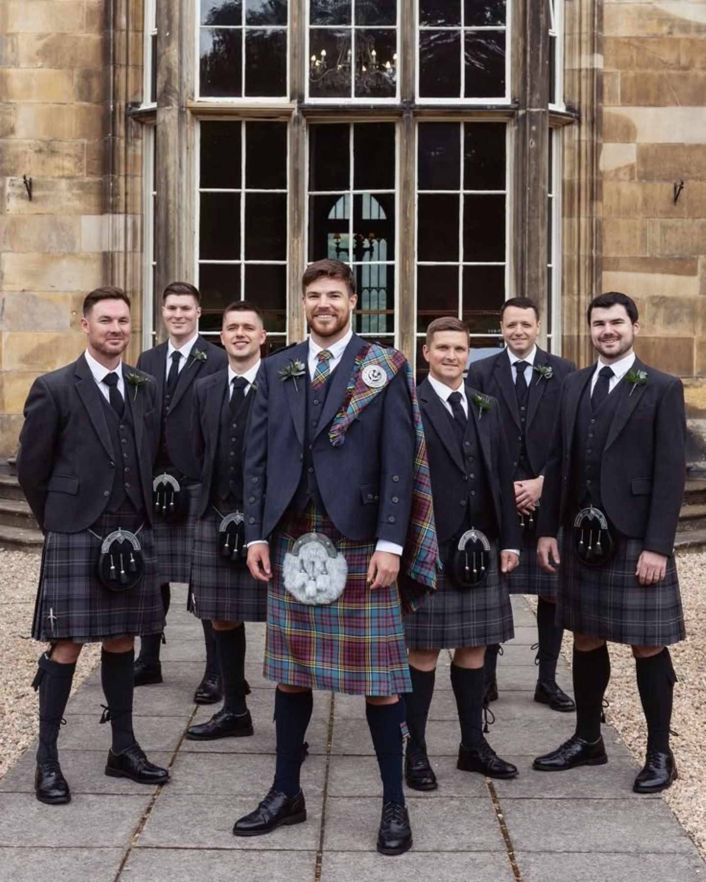 Group of men in formal attire with kilts standing in front of a stone building.